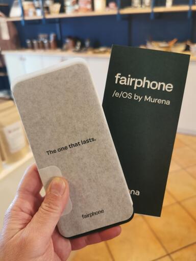 A hand holds a sleek, minimalist fairphone device, showcasing its distinctive design and eco-conscious branding. The phone’s back panel, adorned with the phrase 'The one that lasts,' reflects the brand’s commitment to sustainability and durability. Beside it, the black packaging proudly displays 'fairphone /e/OS by Murena,' emphasizing its ethical and open-source approach.