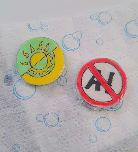 Two pins I made, one is showing a solarpunk symbol (yellow and green flag separated diagonally, like anarchist flags, with a half of a sun symbol, representing solar power, on the green side and a half of a gear symbol on the yellow side, representing technology) and the other one has "AI" written on it, crossed out with a red stroke