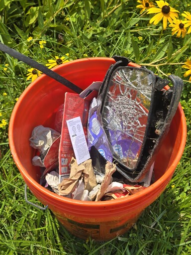 A 5 gallon orange bucket on grass filled with garbage
