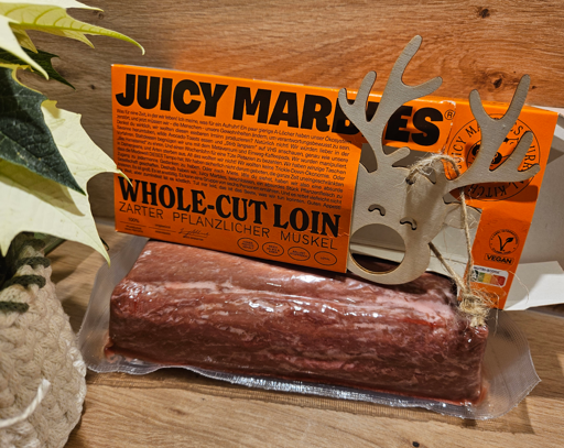 Das eingeschweißte "Whole-Cut Loin" der Firma "Juicy Marbles" auf einem Küchentisch. Darüber liegt die orangefarbene Verpackung mit schwarzem Text, der im Post zitiert ist.